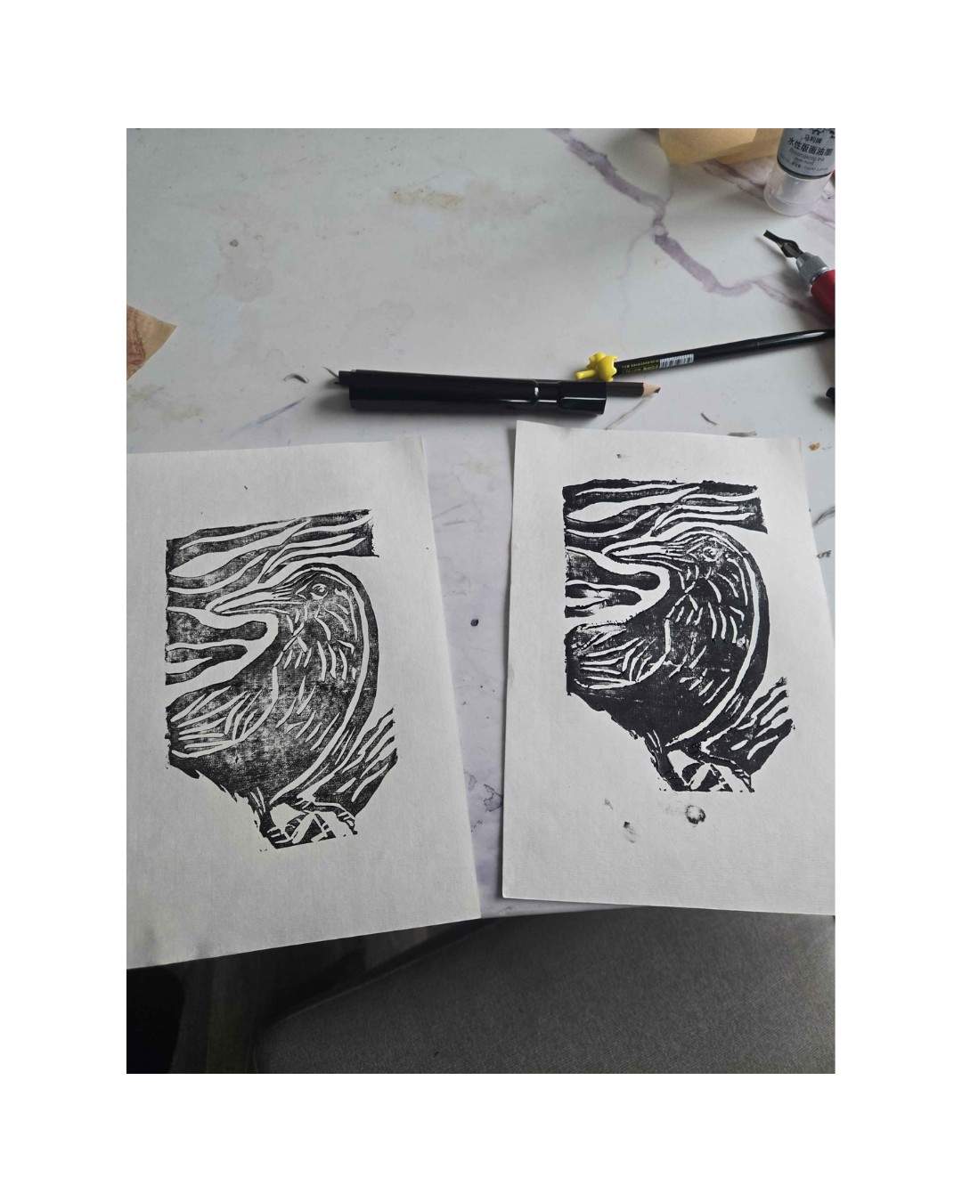 Two black linocut impressions of curling waves lying on a work surface beside printmaking tools.