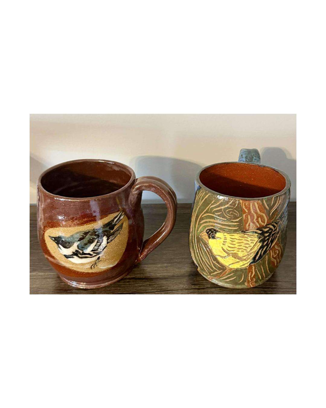 Two glazed mugs, one brown with a black-and-white bird and one green with a yellow bird among branches.